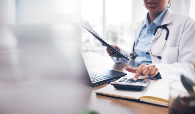 A doctor in a white coat is using a calculator while working on a laptop, highlighting the administrative and financial aspects of medical practice.