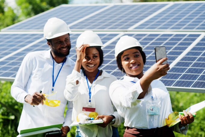 Three engineers, wearing white shirts and hard hats, take a selfie in front of solar panels, smiling and holding work materials, showcasing their involvement in solar energy projects.