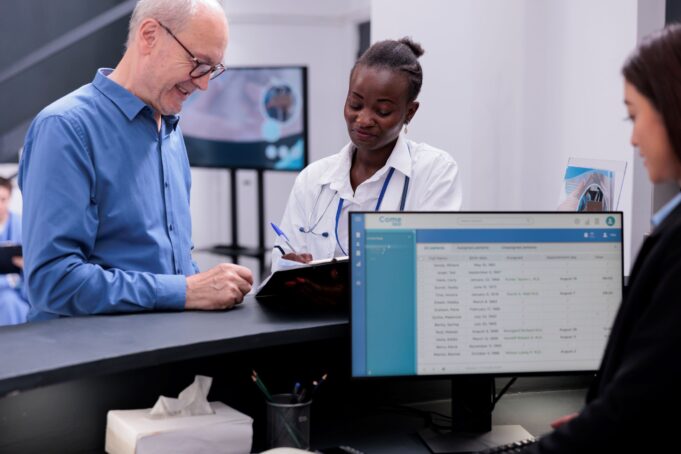 A healthcare professional assists a smiling elderly patient at a reception desk while another staff member works on a computer in a modern medical facility.