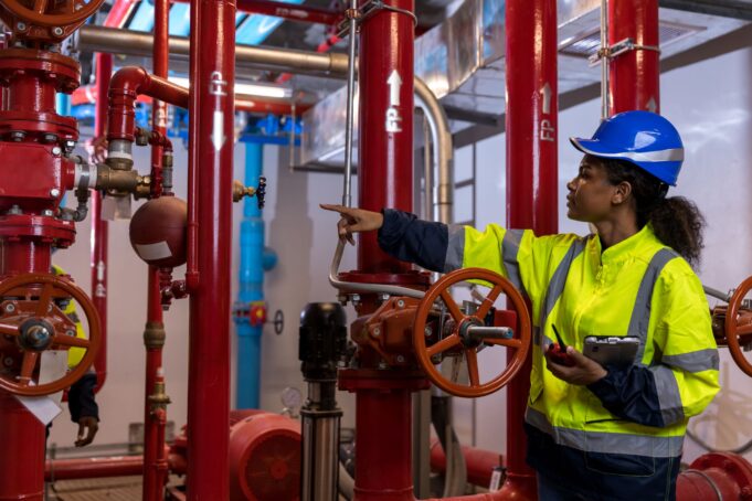 An engineer or technician in a high-visibility jacket and blue hard hat is inspecting a complex system of red industrial pipes and valves inside a facility.